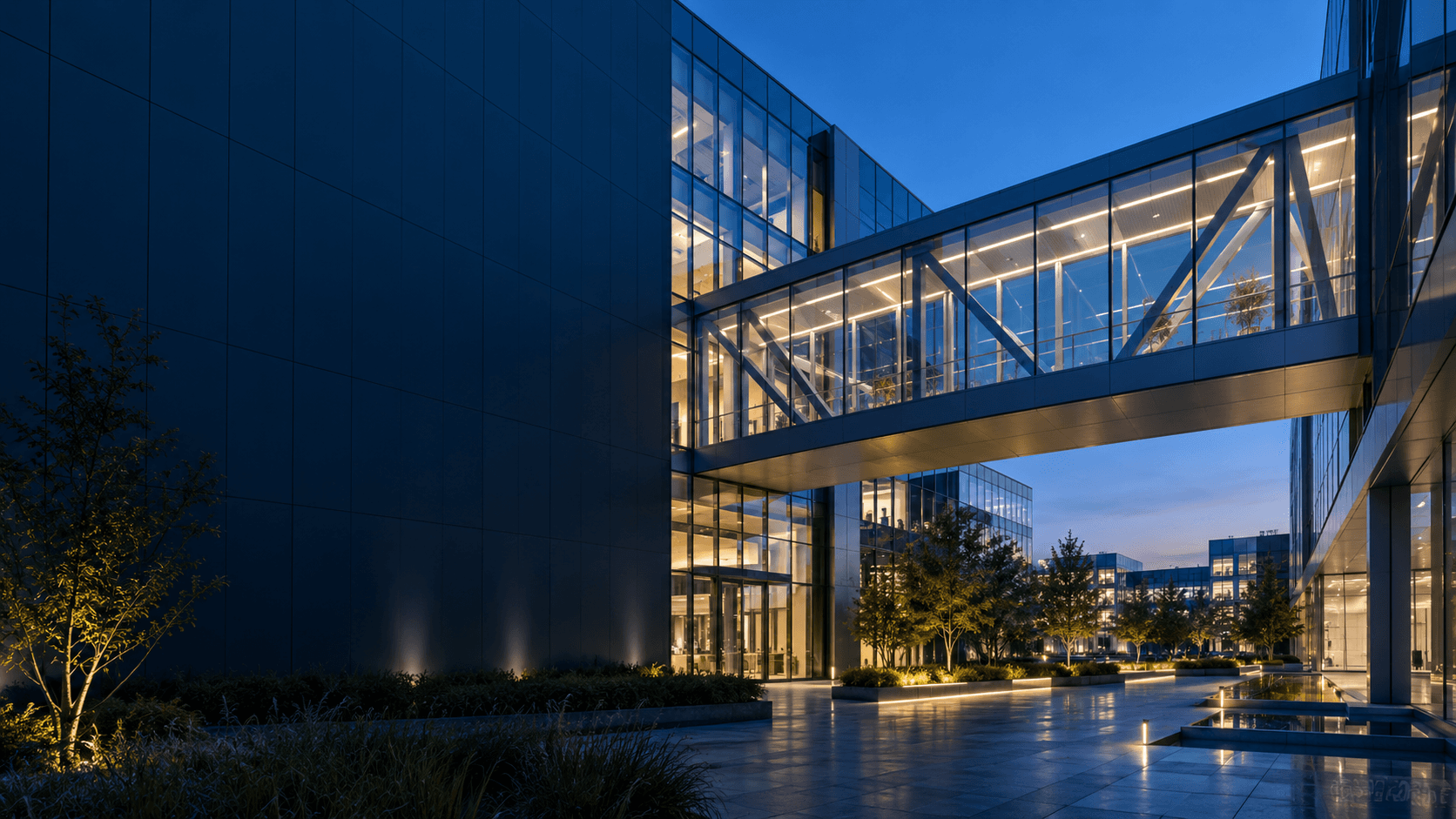 A glass walkway connects two contemporary buildings at blue hour, with cool blue and warm interior lighting